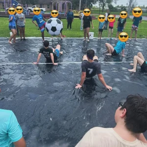 A group of children and young adults, with their faces covered by sunglasses emojis, are enjoying an outdoor slip-and-slide event on a dark tarp, with some playing in the water and others standing around a giant soccer ball and a playground in the background.