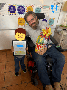 A smiling child holding a Super Star of the Month certificate stands beside an adult staff member in a wheelchair holding a gift basket, celebrating the child’s growth, kindness, and school success in a warm, supportive foster care setting.