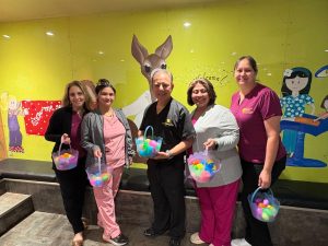 Dr. Joey Cazares and four team members from McAllen Dental Associates hold colorful Easter baskets filled with plastic eggs in front of a cheerful mural — a Sunny Glen donor spotlight moment.