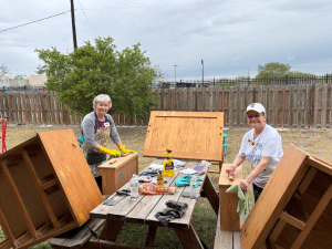 Two Sojourner volunteers — retired Christians who travel across the U.S. and Canada serving ministries — smile while cleaning and restoring wooden furniture at Sunny Glen Children's Home, illustrating the real-world difference in the volunteering vs donating conversation.